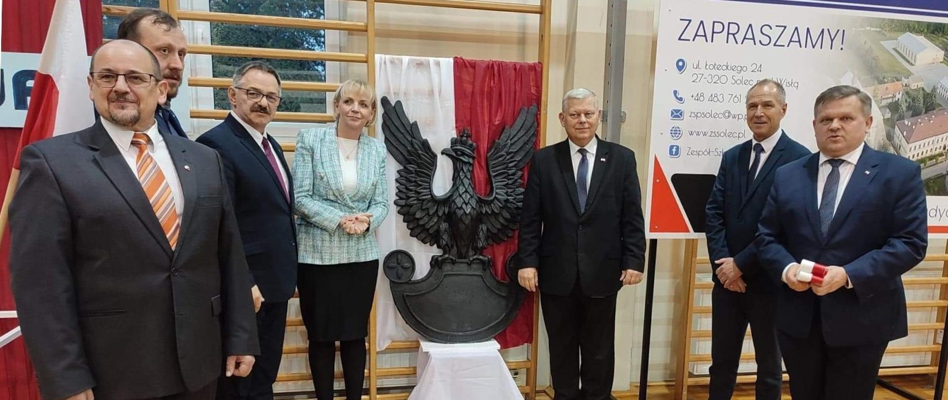 Zaproszeni goście razem z dyrektorem szkoły pozują do zdjęcia z płaskorzeźbą orła legionowego.