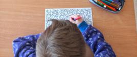 Na zdjęciu widać zdjęcie dziecka robione od góry, tak, że widać że dziecko siedzi przy stoliku, jednak widoczny jest tylko czubek jego głowy. Przed nim jest ławka, na której leży kartka. Dziecko koloruje ją pisakami. Obok kartki leży piórnik.