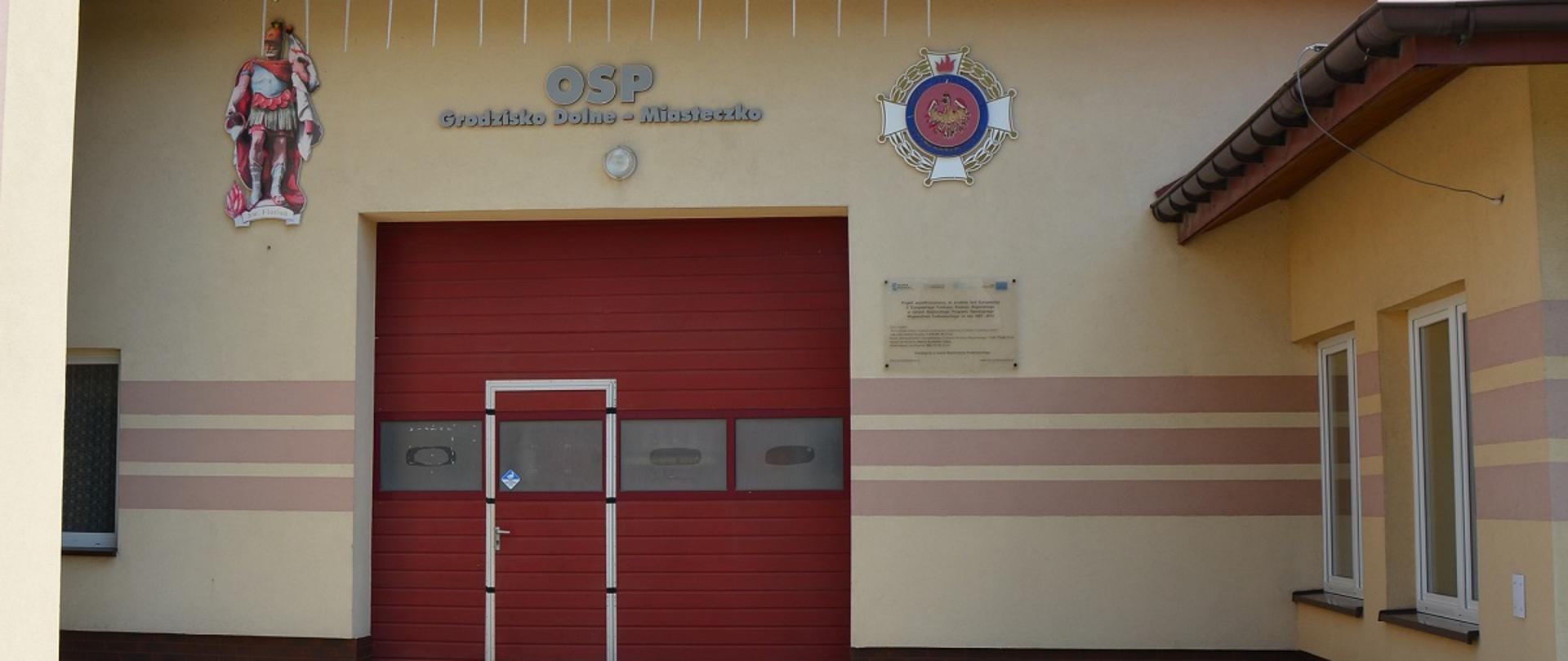 budynek remisy OSP beżowy budynek z czerwonymi dużymi drzwiami garażowymi 
