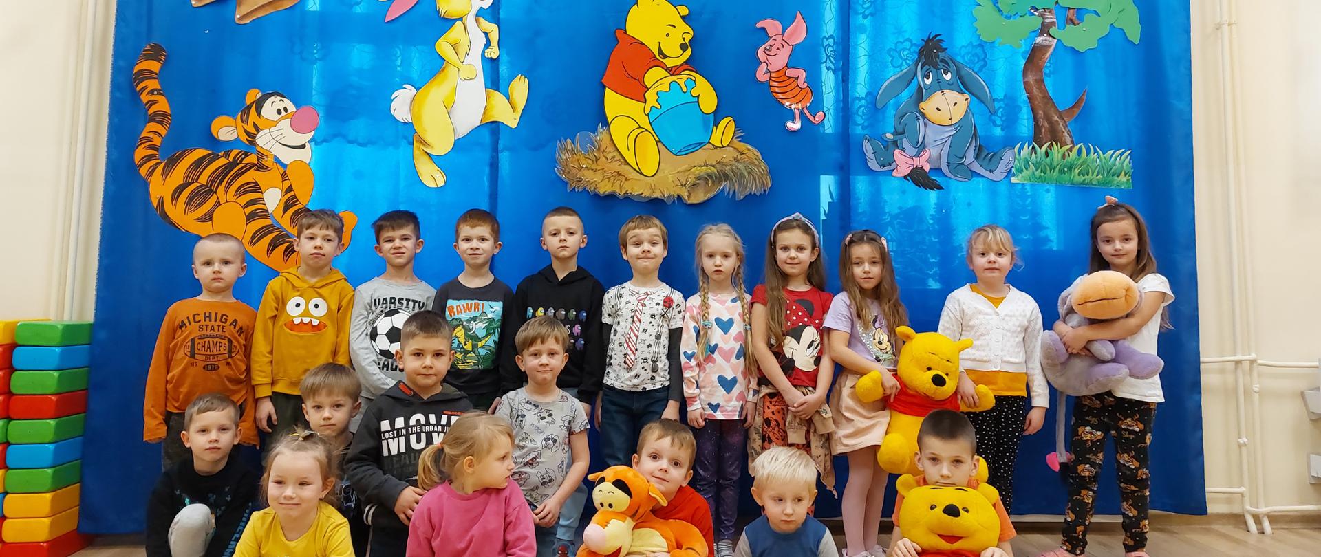 Grupowe zdjęcie przedszkolaków na tle dekoracji z przyniesionymi maskotkami Kubusia Puchatka, Tygryska oraz Kłapouchego. 
