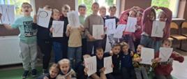 Grupowe zdjęcie uczniów szkoły podstawowej, stojących i siedzących na zielonym dywanie. Dzieci trzymają w rękach białe kartki z pojedynczymi literami, które razem tworzą hasło: " W przyjaźni moc"