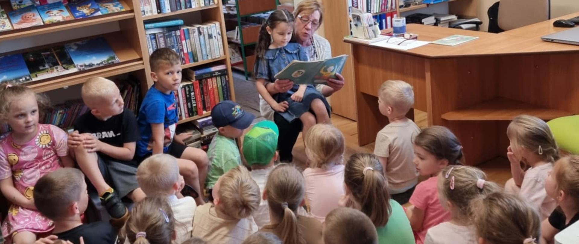 Grupa dzieci w bibliotece słucha czytanej książki przez panią bibliotekarkę z okazji Ogólnopolskiego Tygodnia Czytania