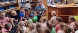 Grupa dzieci w bibliotece słucha czytanej książki przez panią bibliotekarkę z okazji Ogólnopolskiego Tygodnia Czytania