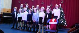 Na tle świątecznej scenografii grupa dzieci z gwiazdą na głowach śpiewa kolędę