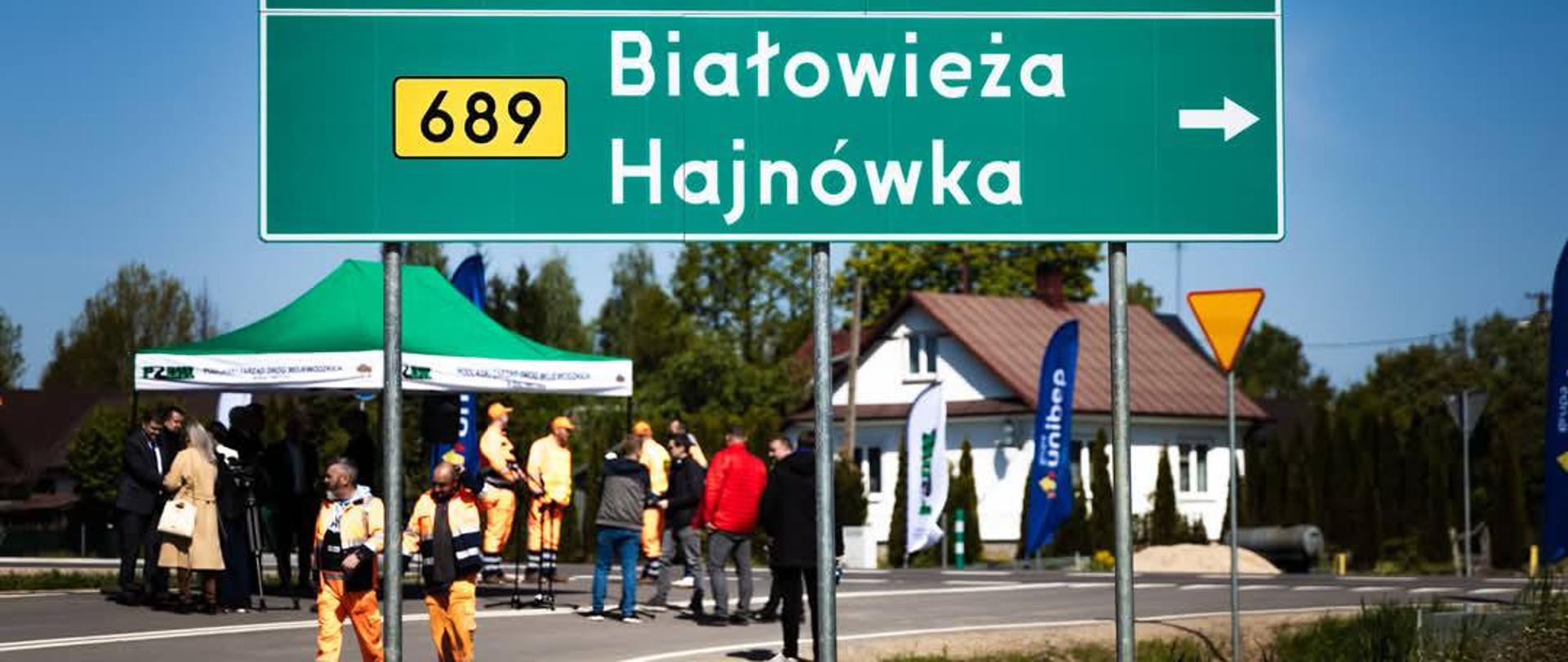 Za znakiem drogowym, informującym o kierunku miejscowości, widać uczestników wydarzenia