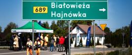 Za znakiem drogowym, informującym o kierunku miejscowości, widać uczestników wydarzenia