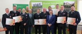 Zdjęcie grupowe wykonane na tle ścianki promocyjnej Powiatu Sokołowskiego. Na zdjęciu przedstawiciele jednostek OSP prezentujący promesy z przyznanymi dofinansowaniami oraz organizatorzy i goście.
