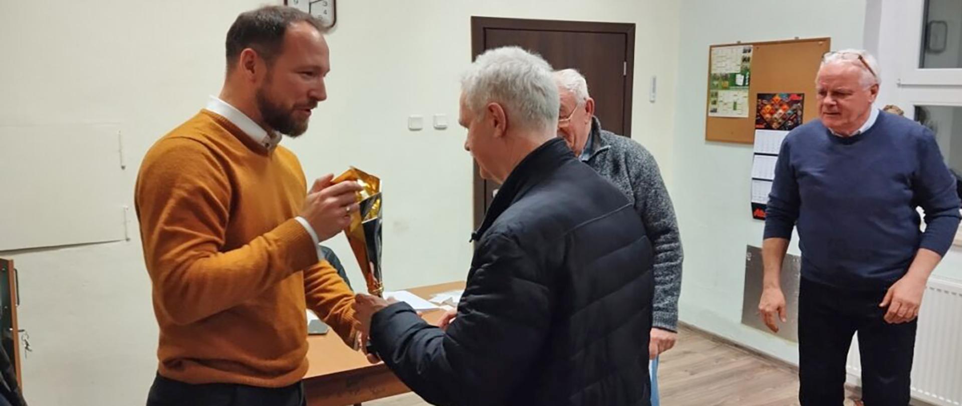 Mężczyzna w pomarańczowym swetrze wręcza złoty puchar starszemu mężczyźnie. W tle dwóch innych mężczyzn. Wszyscy znajdują się w jasnym pomieszczeniu.