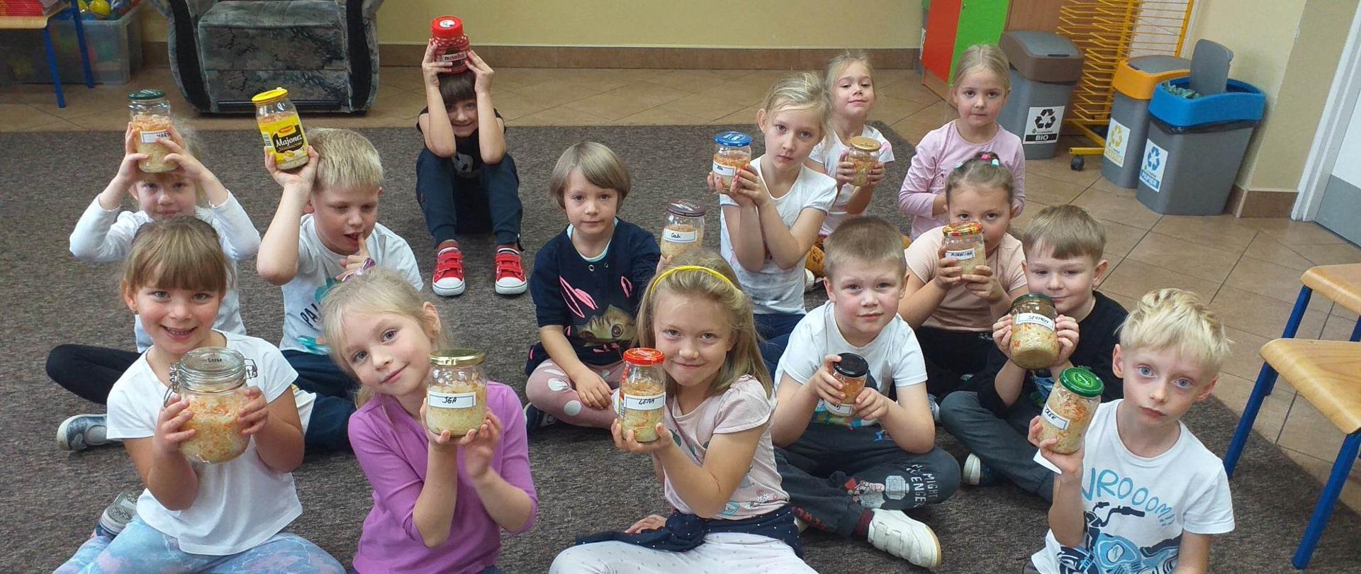 Dzieci siedzące na podłodze. Trzymają w rękach słoiki