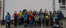 Uczniowie naszej szkoły pozują w strojach narciarskich przed udziałem w Zawodach narciarskich o Puchar Wójta Gminy Łabowa, w tle szara ściana szkoły podstawowej w Nowej Wsi z herbem gminy Łabowa
