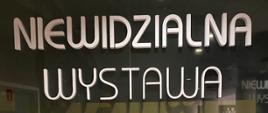 Wejście do muzeum z napisem "Niewidzialna Wystawa" (Niewidzialna Wystawa) w dwóch rzędach białych, dużych liter. Pod tekstem znajduje się ten sam tytuł zapisany alfabetem Braille'a za pomocą białych, wypukłych punktów.