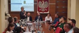 Osoby siedzące przy drewnianym stole. Jedna z nich przemawia do mikrofonu. W tle drewniana ściana oraz biały orzeł w koronie na czerwonym tle.