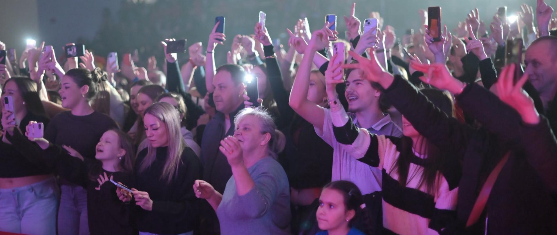 Tłum ludzi podczas koncertu, stojących przy barierce; wiele osób trzyma w górze telefony i nagrywa występ, wśród publiczności widać dorosłych i dzieci, oświetlonych kolorowymi światłami sceny.