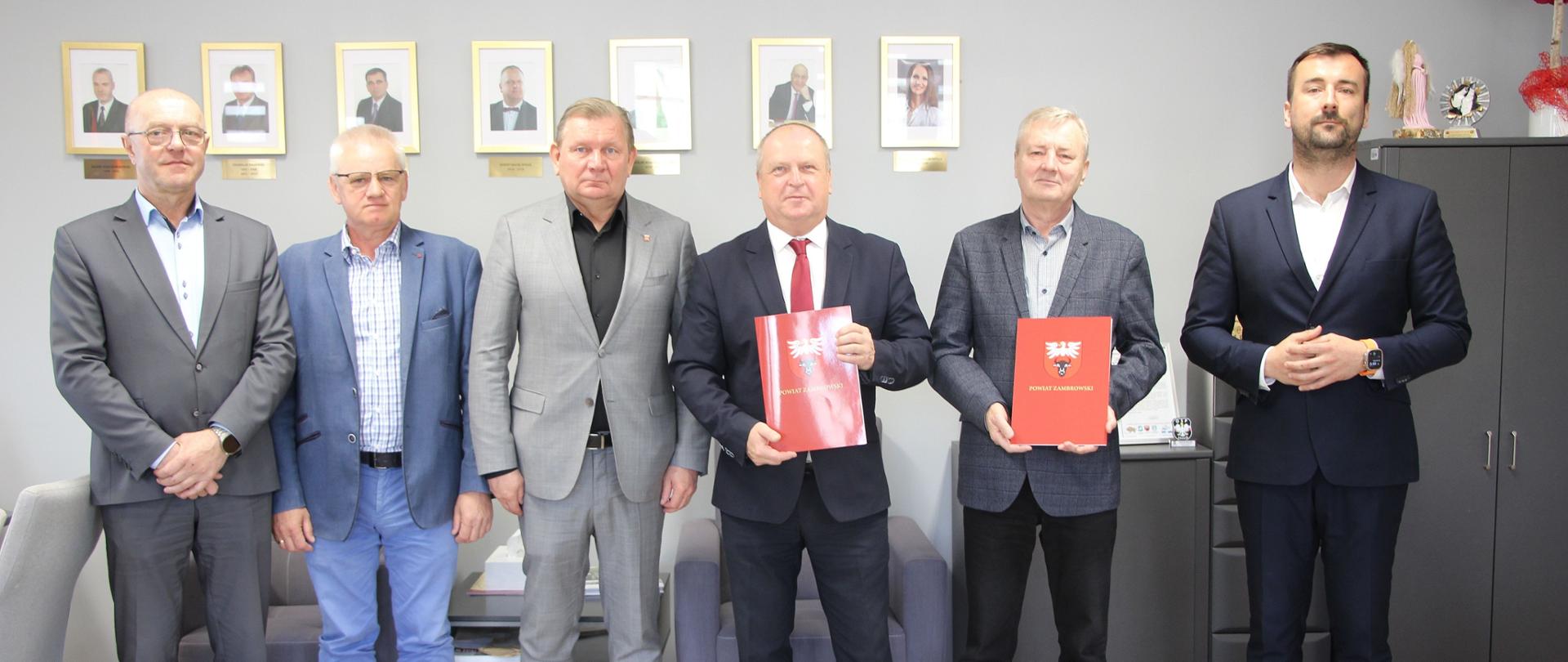 Zdjęcie przedstawia grupę sześciu mężczyzn w garniturach, stojących w rzędzie w oficjalnym pomieszczeniu biurowym. Dwóch z nich trzyma czerwone teczki z godłem