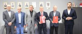 Zdjęcie przedstawia grupę sześciu mężczyzn w garniturach, stojących w rzędzie w oficjalnym pomieszczeniu biurowym. Dwóch z nich trzyma czerwone teczki z godłem