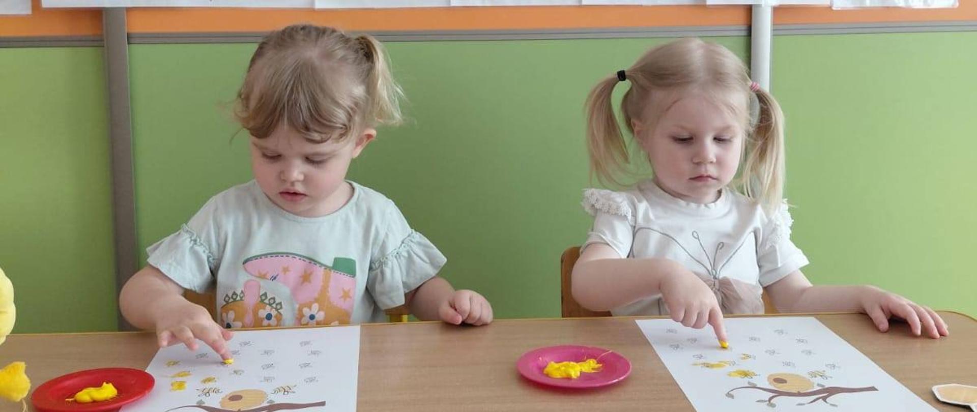 Na zdjęciu są dwie dziewczynki z grupy Motylki.i. Za nimi widnieje napis " Dzień Pszczoły". Dziewczynki wykonują pracę plastyczną farbami.