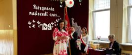 Pani wójt przemawia na zakończenie roku szkolnego, za nią poczet sztandarowy, na ścianie na bordowej kotarze umieszczona dekoracja w postaci motylków unoszących się znad otwartej książki i napis "Pożegnania nadszedł czas..."