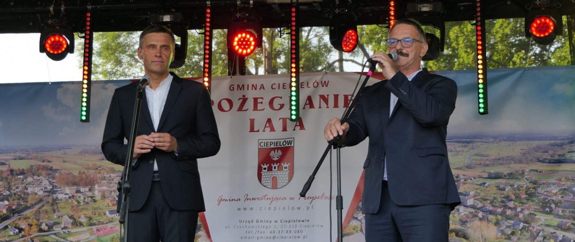Wójt Gminy Ciepielów oraz Starosta Lipski witają zaproszonych gości. Za nimi baner wydarzenia, nazwą Pożegnanie lata, herbem Gminy Ciepielów i panoramą Ciepielowa z lotu ptaka.