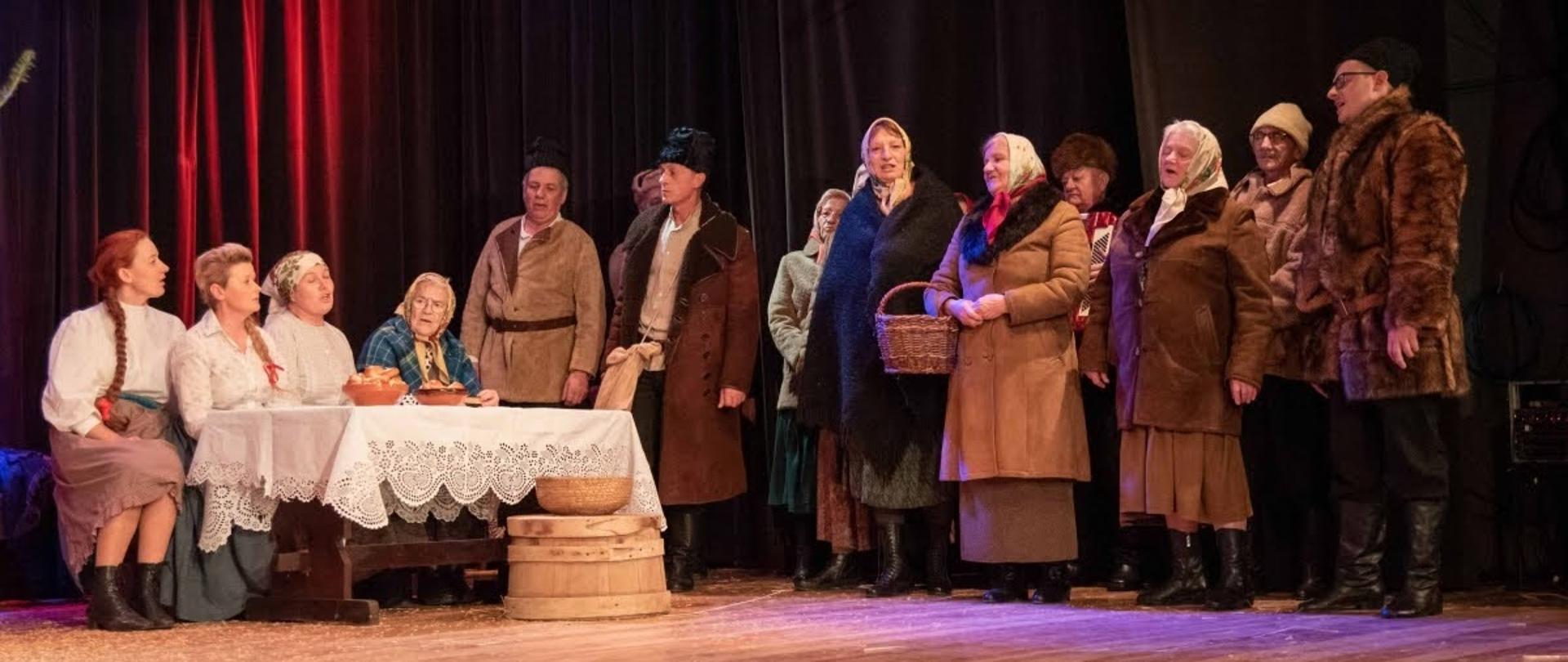 aktorzy ubrani w stroje ludowe występują na scenie, cztery kobiety siedzą przy stole reszta stoi 