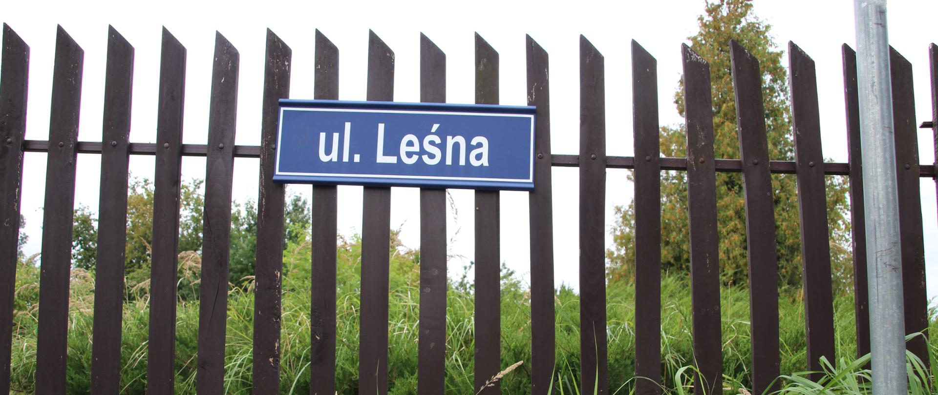 Zbliżenie na niebieską tabliczkę z napisem "ul. Leśna" umieszczoną na tle ciemnobrązowego, drewnianego płotu ze szpiczastymi sztachetami. Na pierwszym planie widać wysoką, zieloną trawę.