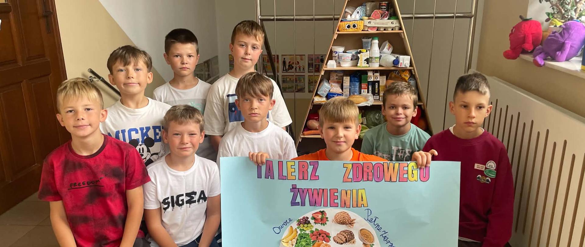 Uczniowie klasy 3a prezentują wykonany plakat „Talerz zdrowego żywienia” na tle szkolnej piramidy zdrowego żywienia.