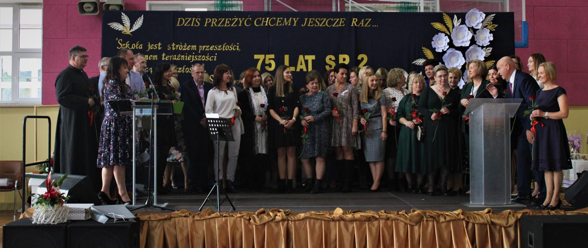 Zdjęcie przedstawia kadrę nauczycielską Szkoły Podstawowej numer 2 w Przasnyszu zgromadzoną na scenie.