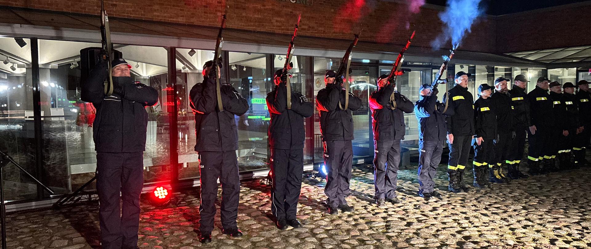 Na tle przeszkolonego budynku w szeregu stoją umundurowani policjanci i strażacy. Sześciu pierwszych policjantów (od lewej) oddaje salwę honorową z karabinków. 