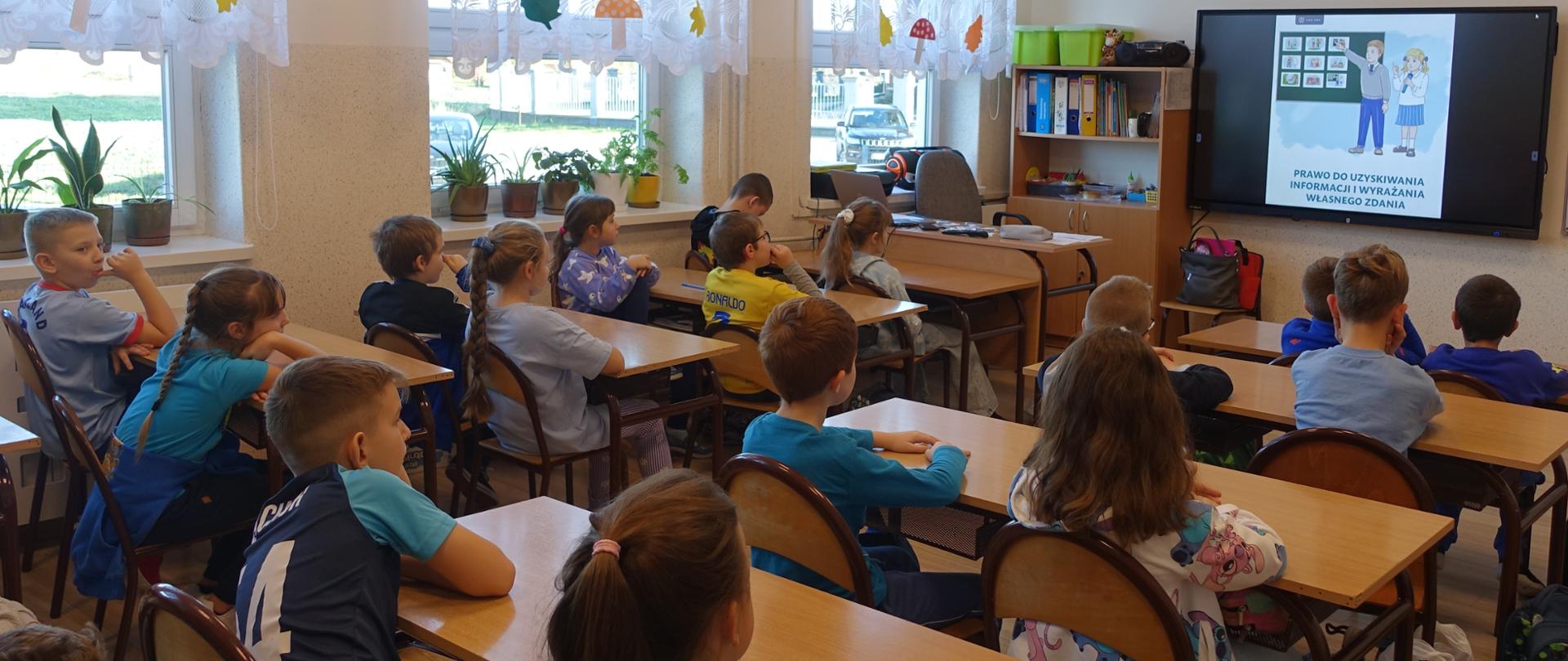 Uczniowie klas I–IV siedzą w ławkach w sali lekcyjnej i wspólnie oglądają film dotyczący praw dziecka