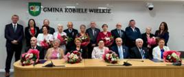 Pary małżeńskie z Gminy Kobiele Wielkie uhonorowane medalami za długoletnie pożycie małżeńskie, stojące i siedzące wspólnie na zdjęciu grupowym, podczas uroczystości. Panie trzymają bukiety kwiatów, a na piersi Jubilatów widoczne są medale. Po lewej stronie Wójt Gminy Beata Pokora oraz Przewodniczący Rady Gminy Adam Kleszczewski, a po prawej Z-ca Kierownika Urzędu Stanu Cywilnego w Kobielach Wielkich Joanna Zakrzewska. W tle na ścianie logo GMINA KOBIELE WIELKIE z herbem gminy.