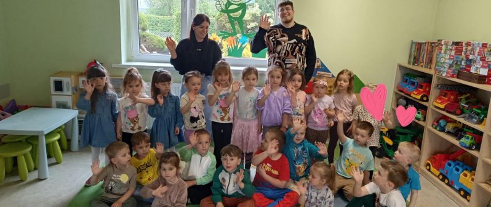 Grupa dzieci w wieku przedszkolnym, dziewczynki stoją w rzędzie, obok siebie, przed nimi na kolorowym dywanie siedzą dziewczynki i chłopcy.Za dziećmi stoi mężczyzna i kobieta. Dorośli i dzieci mają podniesioną jedną rękę i otwartą dłoń. Za nimi okno, na szybie naklejony obraz żaby i kwiaty. Za oknem widać zieleń. Po prawej stronie regały z książkami, zabawkami, puzzlami. Po lewj mały stolik i taborety. ściany w kolorze zielonym. 