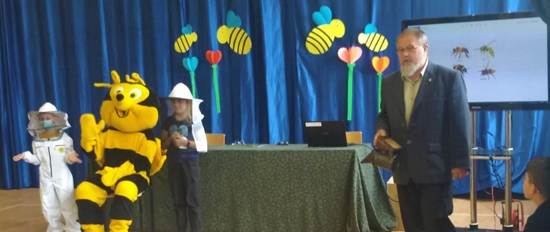 Dwoje dzieci w strojach pszczelarza stoi obok dużej maskotki pszczoły. Po ich lewej stronie stoi mężczyzna w marynarce i przemawia