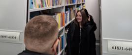Uczniowie zwiedzają bibliotekę, uczennica przechodzi wzdłuż regału z książkami
