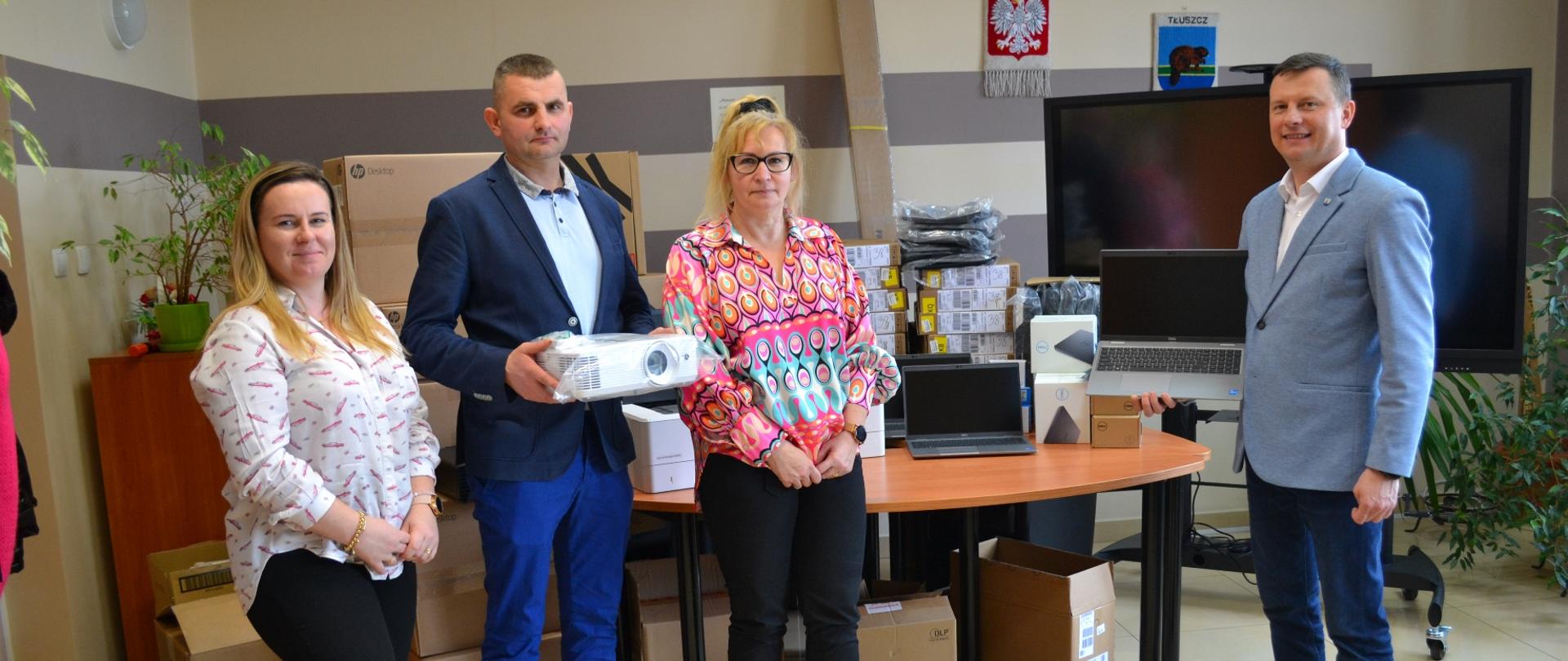 Dwie kobiety oraz dwoje mężczyzn w marynarkach. Za nimi stoi stół z kartonami i laptopem