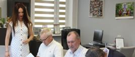 Zdjęcie przedstawia moment podpisywania dokumentów przez mężczyzn siedzących przy długim stole konferencyjnym. Stojąca obok kobieta w białej sukience w grochy przygląda się całemu procesowi.