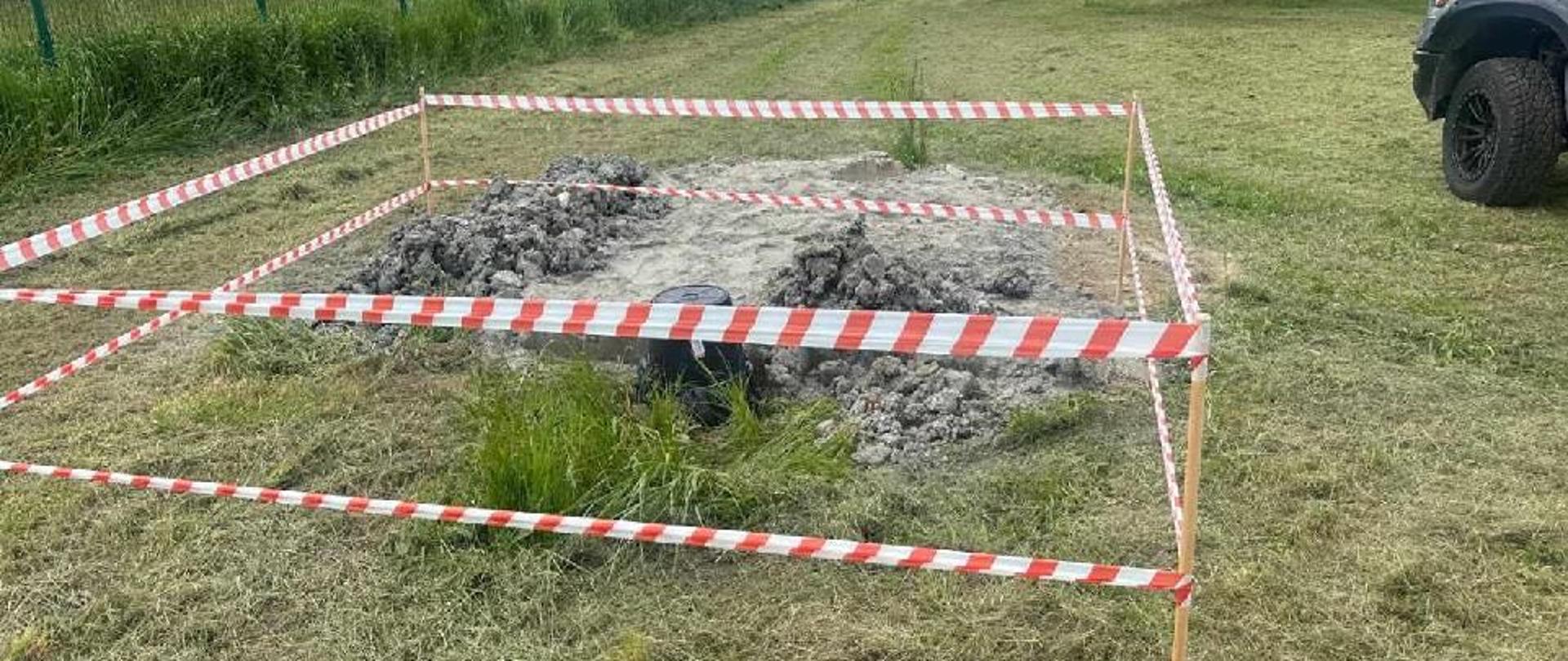 Zdjęcie przedstawia ogrodzony taśmami fragment trawnika, na którym znajduje się studnia głębinowa.