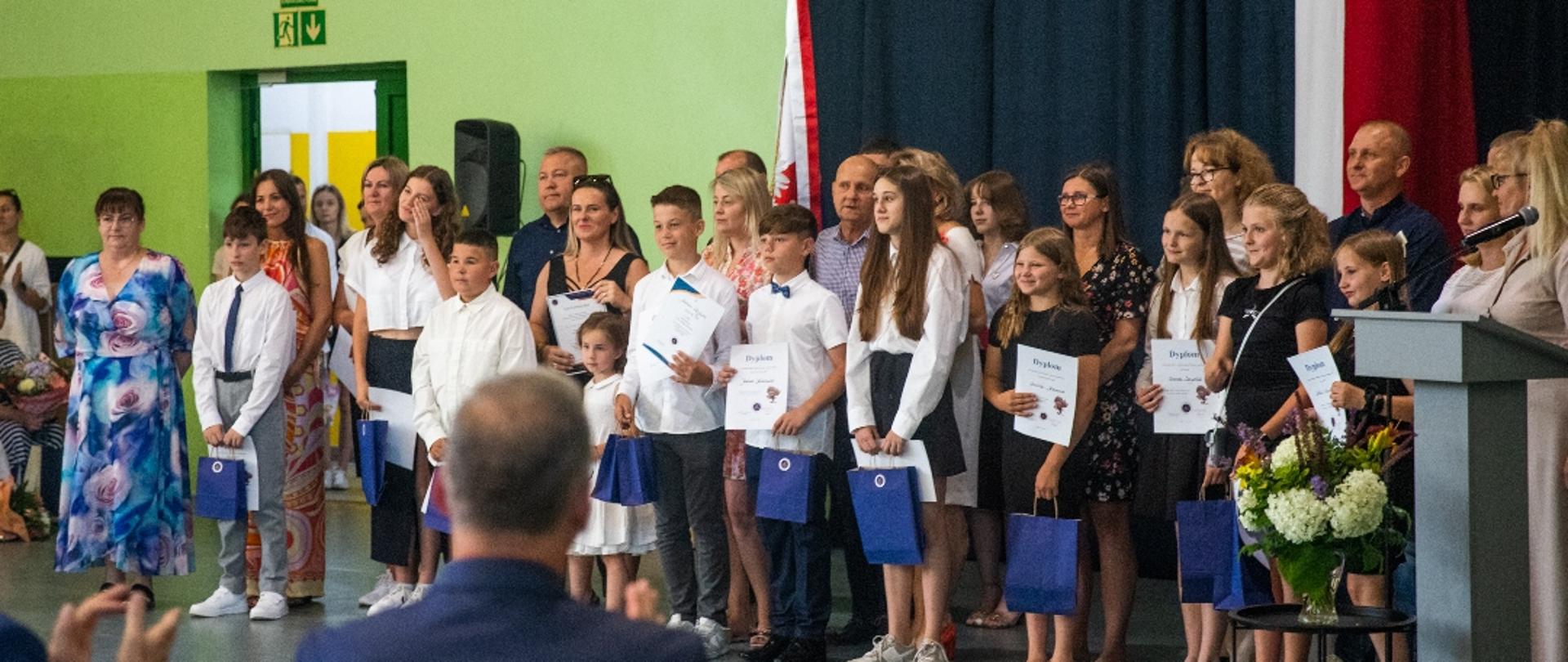 Grupa dzieci stoi w szeregu. Ubrani w większości w białe koszule. W opuszczonych rękach trzymają niebieskie papierowe torebki. 
