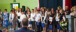 Grupa dzieci stoi w szeregu. Ubrani w większości w białe koszule. W opuszczonych rękach trzymają niebieskie papierowe torebki. 