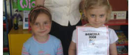 Kobieta pozuje do zdjęcia z dwiema dziewczynkami, które trzymają w rękach zdjęcie grupy i dyplom