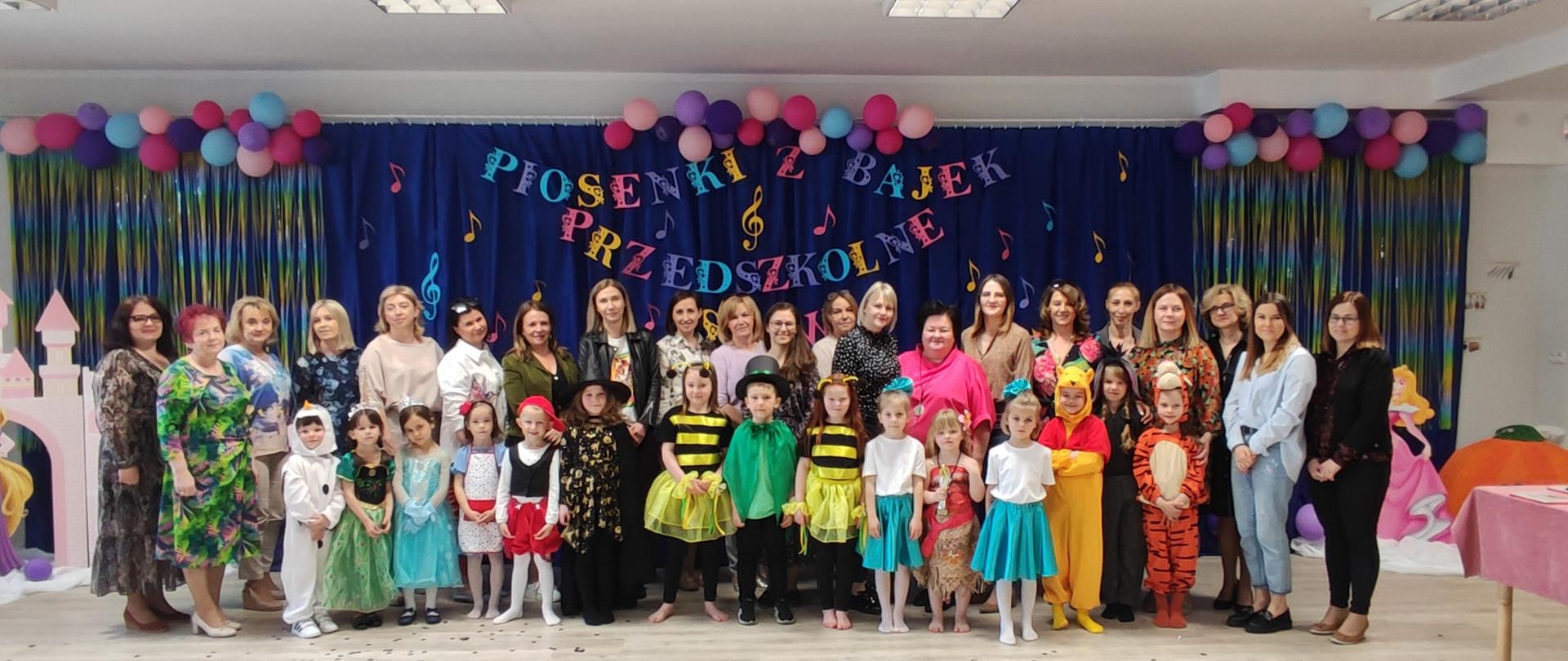 Grupa dzieci i nauczycieli na tle dekoracji z napisem Piosenki z Bajek - Przedszkolne Show