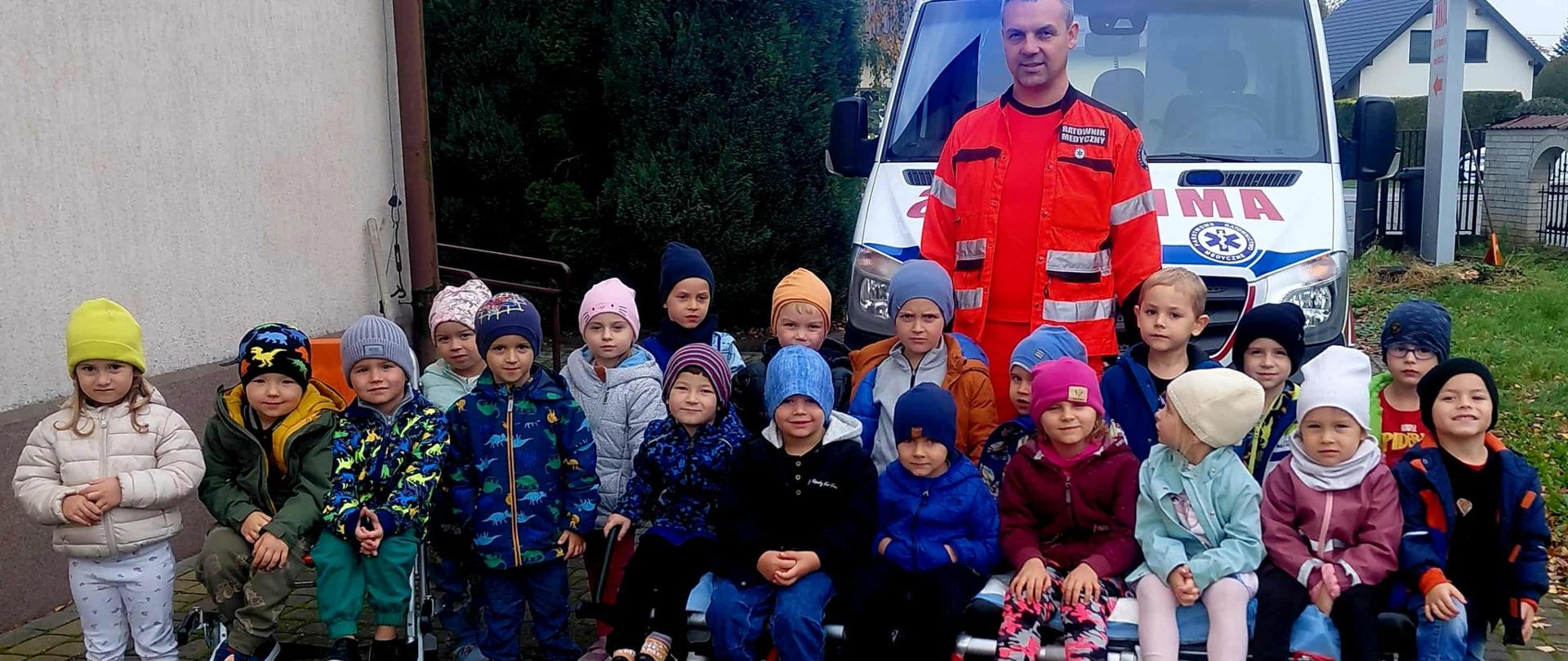 Grupa dzieci pozuje do zdjęcia z ratownikiem medycznym przy karetce.