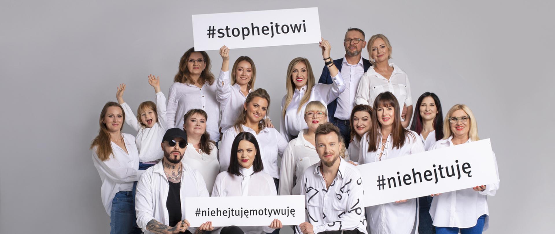 Zdjęcie przedstawia wiele osób w białych koszulach, niebieskich lub czarnych spodniach na szarym tle. W dłoniach trzymają tablice z napisami #stophejtowi #niehejtuję #niehejtujęmotywyję.