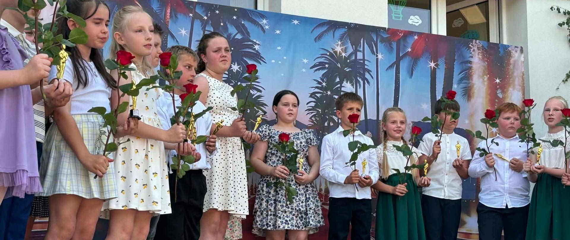 Na zdjęciu widać grupę dzieci stojących na scenie podczas szkolnego występu lub uroczystości. Uczniowie są elegancko ubrani – dziewczynki mają sukienki w jasnych kolorach, a chłopcy białe koszule i ciemne spodnie. Każde dziecko trzyma w dłoni czerwoną różę oraz małą złotą statuetkę, co może świadczyć o wręczeniu nagród lub podziękowań.
W tle znajduje się dekoracyjna ścianka przedstawiająca nocny krajobraz z palmami i gwiazdami, nadająca wydarzeniu uroczysty charakter. Scena ustawiona jest przed budynkiem szkoły, którego okna ozdobiono rysunkami balonów. Dzieci stoją w jednym rzędzie, skupione na swoim występie lub ceremonii, a atmosfera wydarzenia wydaje się podniosła i radosna.