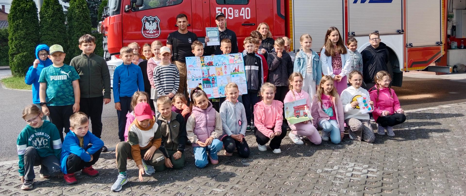 Wizyta w Ochotniczej Straży Pożarnej w Chorzelowie - zdjęcie uczniów ze strażakami na tle samochodu strażackiego.