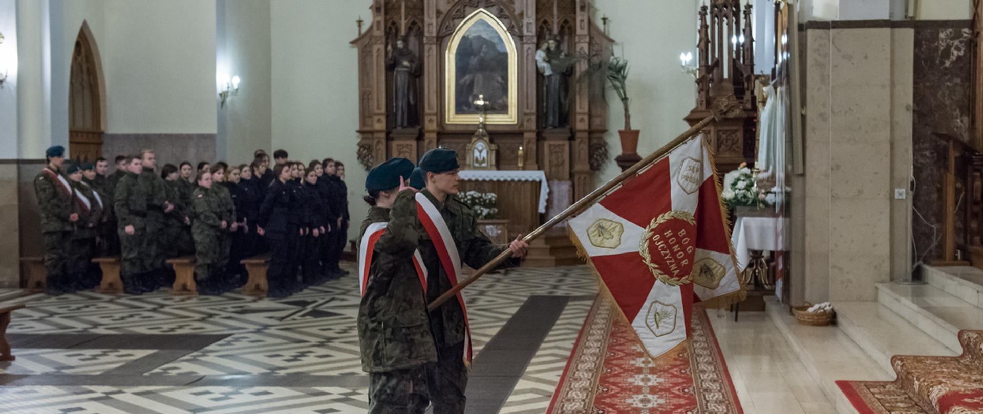 Na zdjęciu uczniowie klas mundurowych ZS nr 1 w Sokołowie Podlaskim prezentujący w kierunku ołtarza sztandar Światowego Związku Żołnierzy Armii Krajowej – Koła w Sokołowie Podlaskim. W tle nawa boczna oraz stojący w mundurach uczniowie.