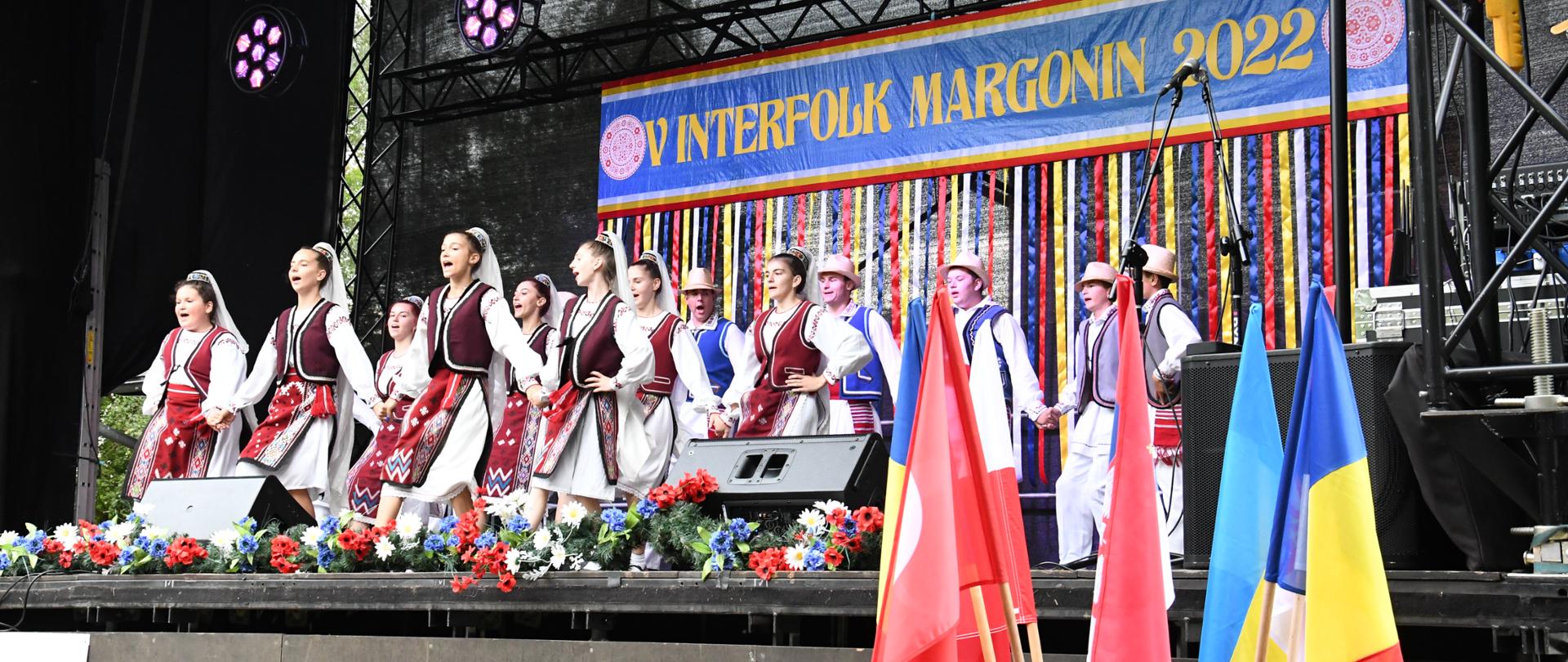 Zespół Doina Casimcei z Rumunii na scenie w komplecie ok. 25 osób, dziewczęta z przodu i chłopcy z tyłu. Ubrani w tradycyjne stroje ludowe. Z tyłu sceny wielki baner z napisem InterFolk Margonin 2022 oraz na pierwszym planie flagi krajów biorących udział w festiwalu.