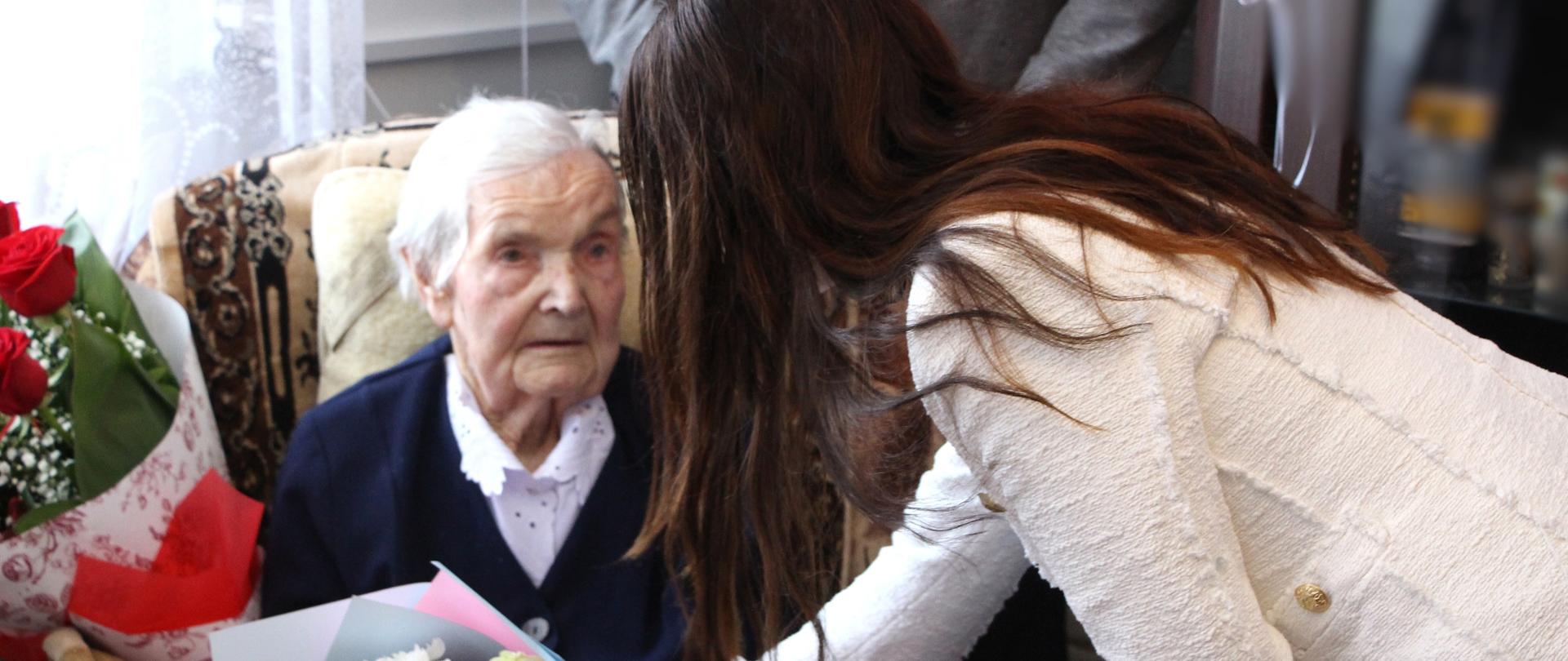 Bliskie ujęcie Pani Aleksandry Orłowskiej, mieszkanki Zambrowa, która 6 lutego 2026 roku ukończyła 100 lat. Jubilatka siedzi w fotelu, trzymając w rękach kolorowy bukiet kwiatów. Nad kobietą pochyla się Edyta Marchelska-Groszfeld (Starosta Powiatu Zambrowskiego), ubrana w jasną, tweedową marynarkę, przekazując jubilatce gratulacje.