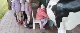 Dziewczynka siedzi na białym krześle i doi sztuczną krowę 