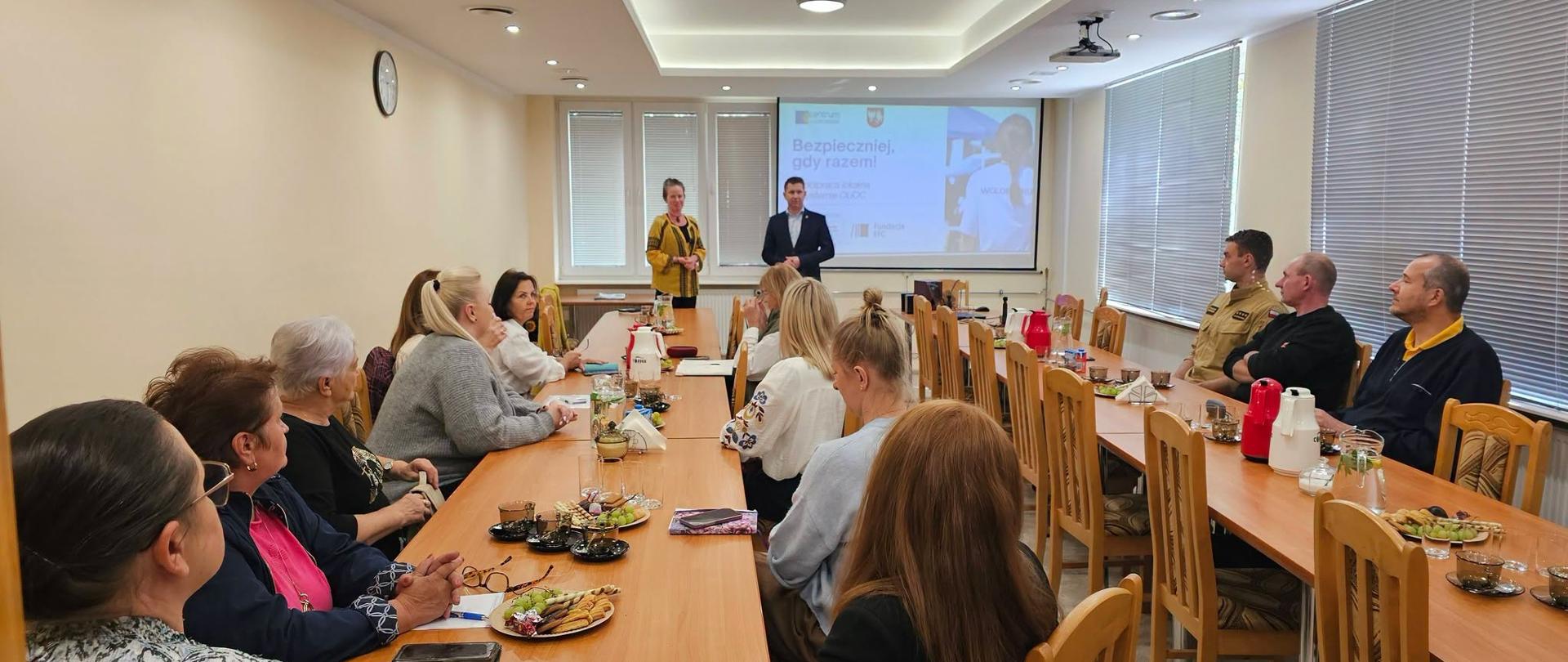 Grupa osób siedzących przy długim stole w sali konferencyjnej, a na ekranie z przodu wyświetlana jest prezentacja.