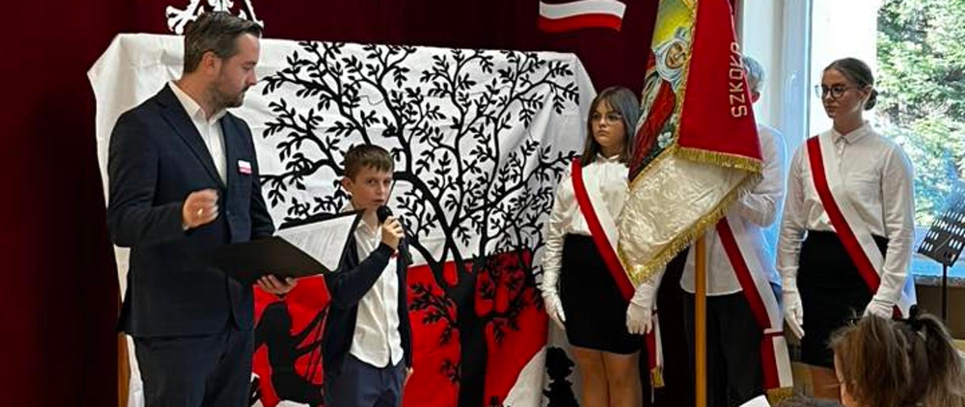 Uczeń przemawia do mikrofonu, obok stoi wychowawca klasy, po lewej stronie poczet sztandarowy ze sztandarem szkoły, w tle dekoracja: na białym płótnie biało-czerwona mapa polski, na niej czarna sylweta drzewa z huśtającą się na huśtawce dziewczynką