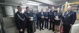 Grupa kobiet i mężczyzn stojących w pomieszczeniu technicznym kotłowni; kilku z nich jest w galowych mundurach strażackich z odznaczeniami, inni w strojach formalnych. Dwie osoby trzymają pamiątkowe upominki. W tle widoczne są instalacje grzewcze oraz baner z napisem „Małopolska”.